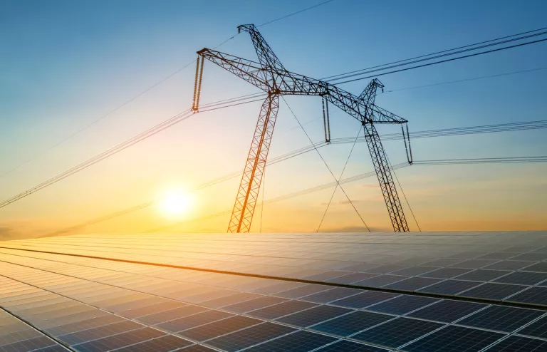 Im Vordergrund ist eine Solaranlage und im Hintergrund ist ein öffentlicher Hochspannungsmast und am Horizont ist der Sonnenaufgang zu sehen.
